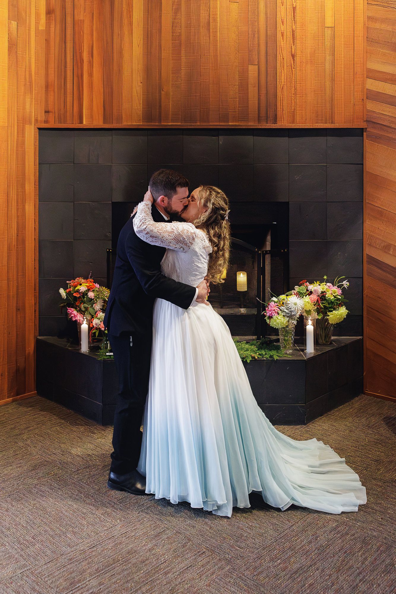 A romantic wedding couple shares an intimate moment in front of a modern fireplace with wood paneled walls.