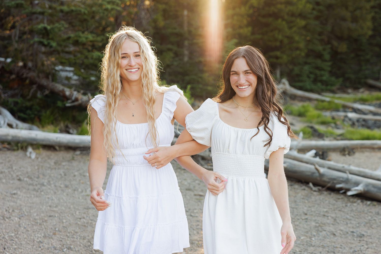 Two young women in white dresses smiling outdoors with sunlight and driftwood in the background.