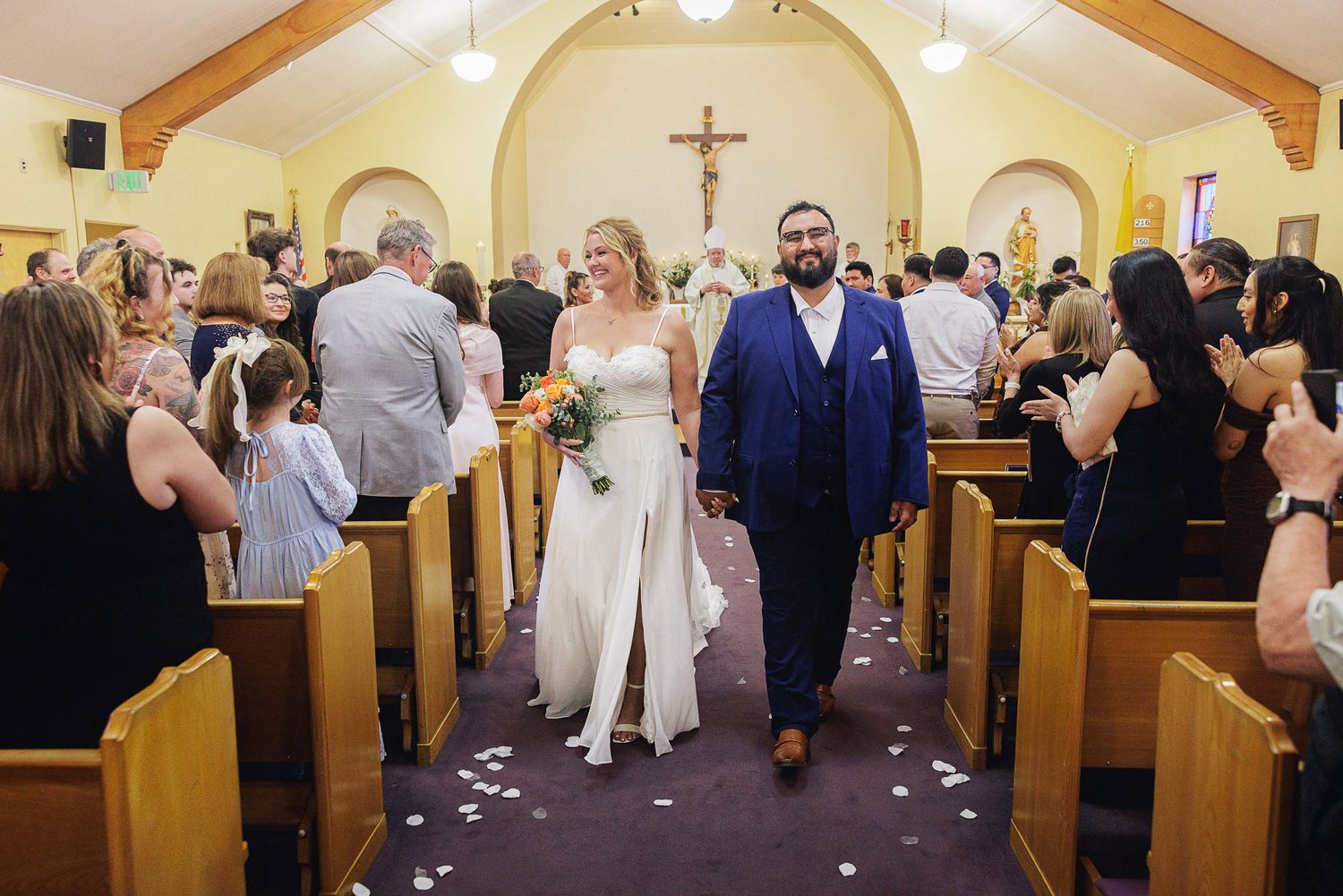 Bride and groom walk down church aisle after wedding ceremony, surrounded by cheering guests throwing flower petals.