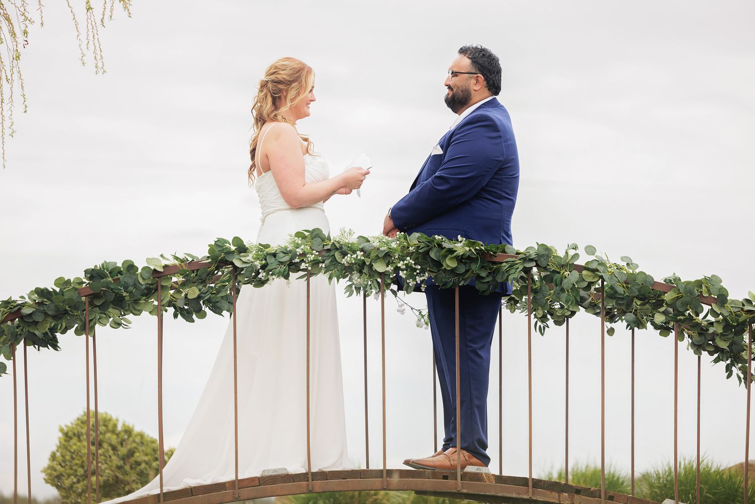 Bride and groom exchange vows on a decorated bridge with lush greenery garland under an overcast sky.