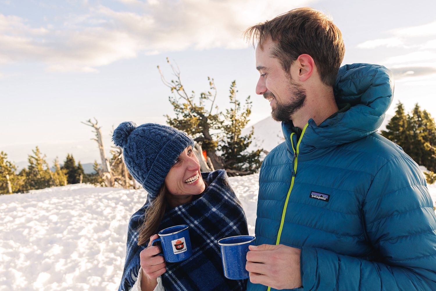 Couple smiling at each other holding blue mugs in a snowy winter landscape wearing warm jackets and a knit hat.