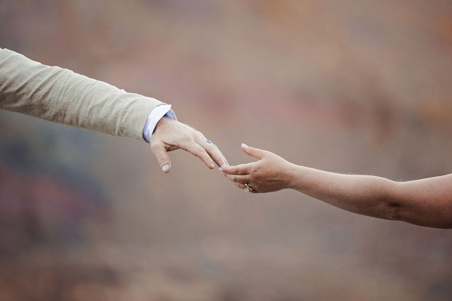 Two hands reaching toward each other against a soft blurred earth-toned background, one in a tan sleeve and the other bare with a ring.