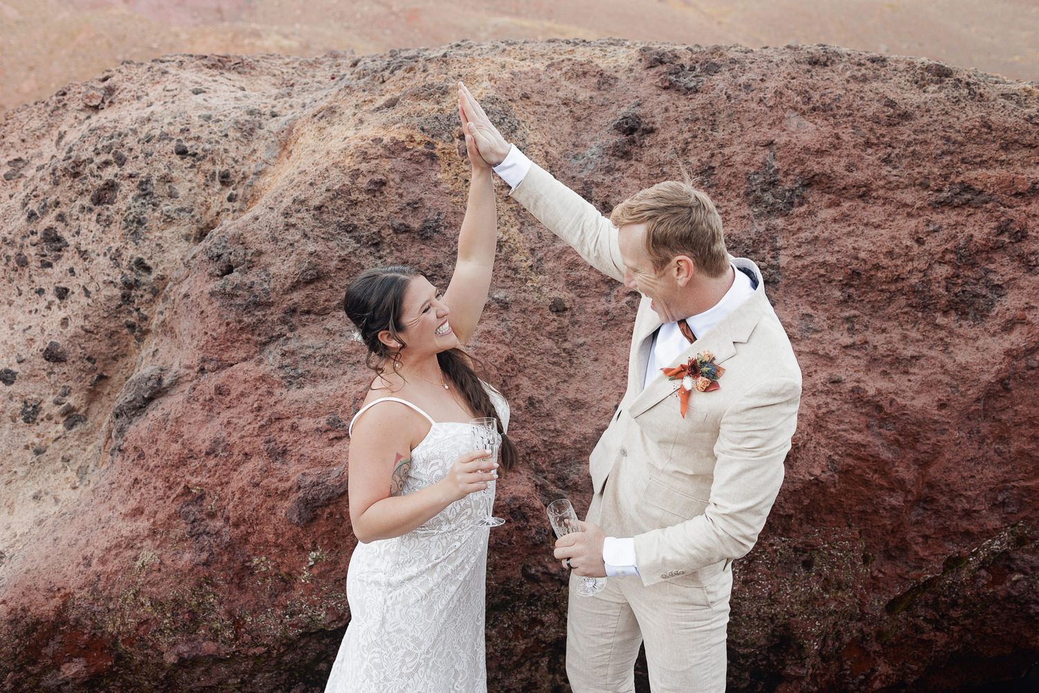 Smiling bride in a white dress twirled by her groom in a cream suit with a floral boutonniere against red volcanic rock.
