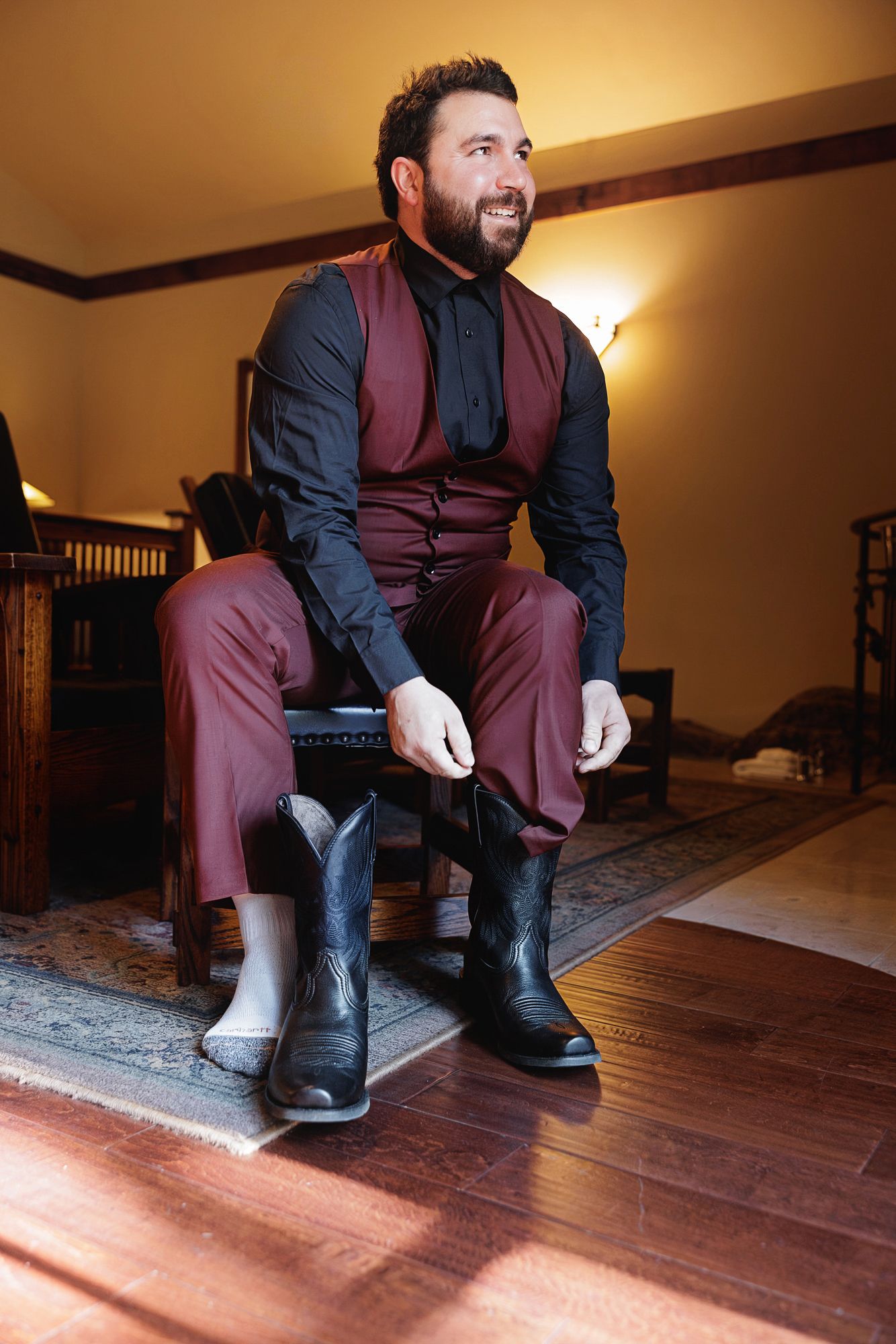 A person in formal attire with a burgundy vest and black boots sits on a wooden chair in a warmly lit room.