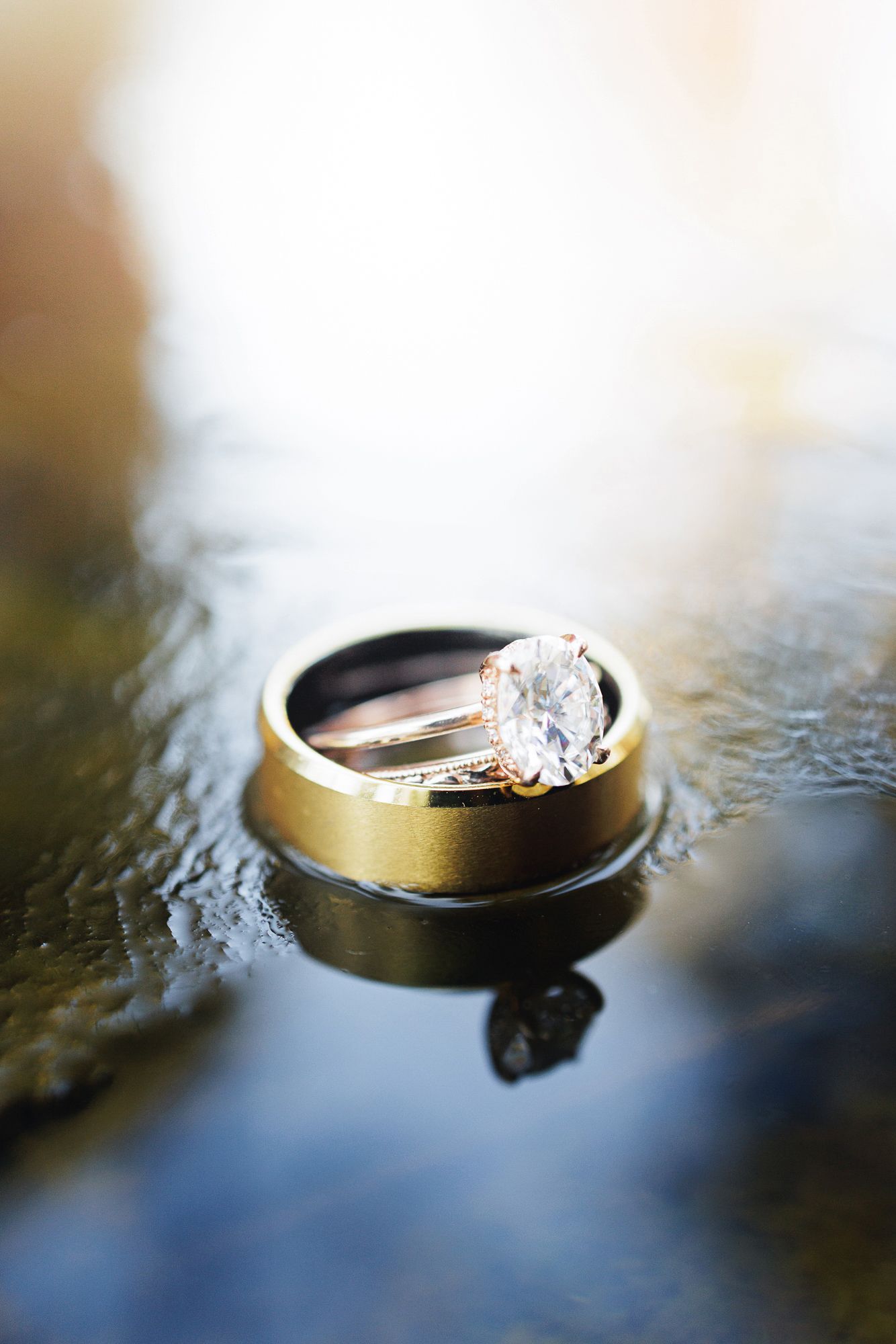 Wedding rings photographed on reflective surface with gold band and diamond engagement ring in artistic macro shot.