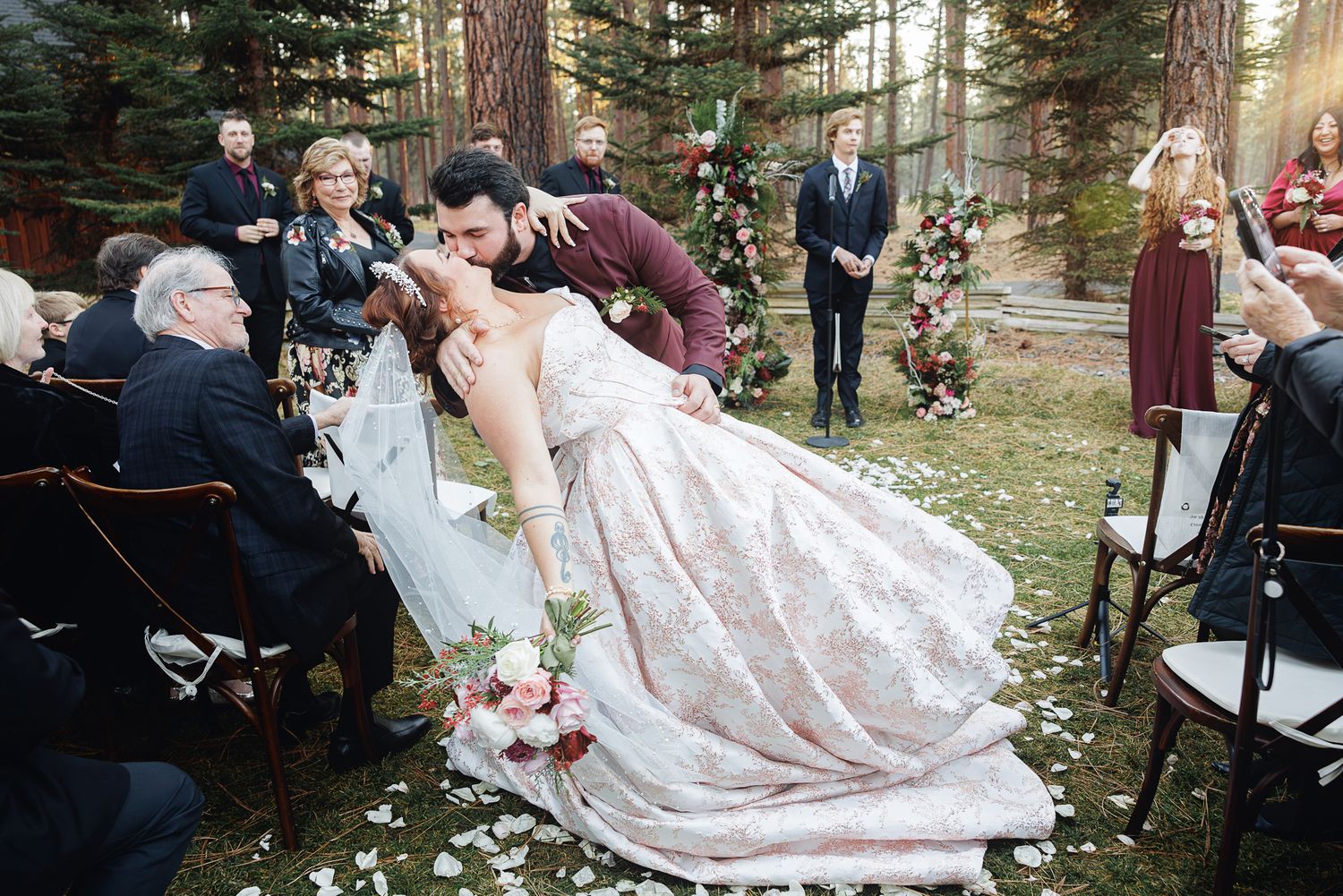 A couple shares a romantic dip and kiss during their outdoor forest wedding ceremony as guests look on.