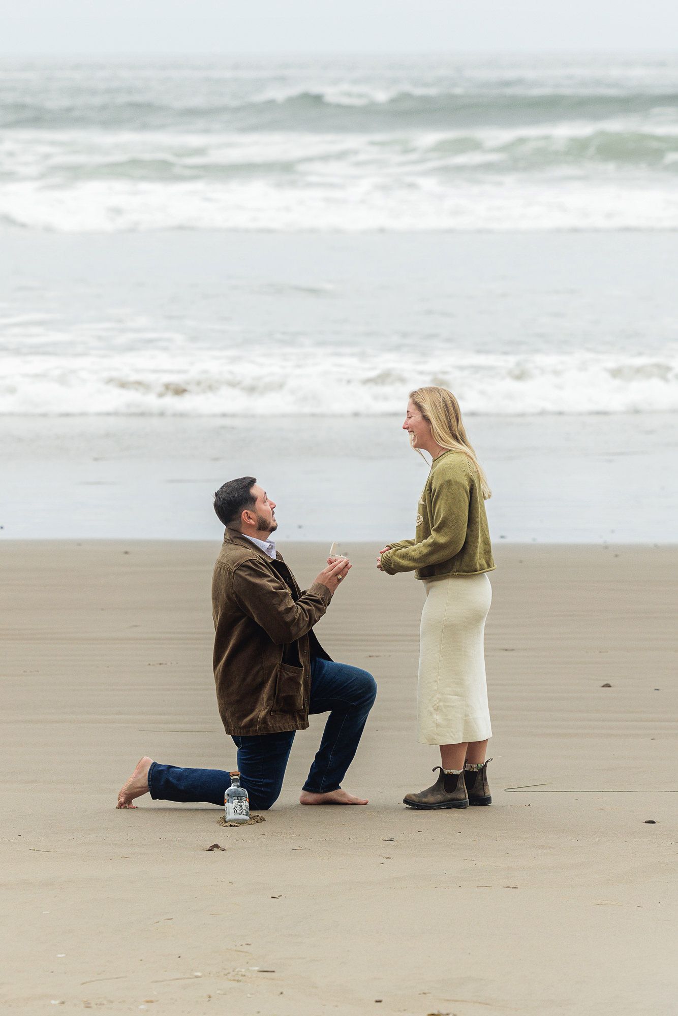 Man proposing on one knee to woman on sandy beach with ocean waves in background.