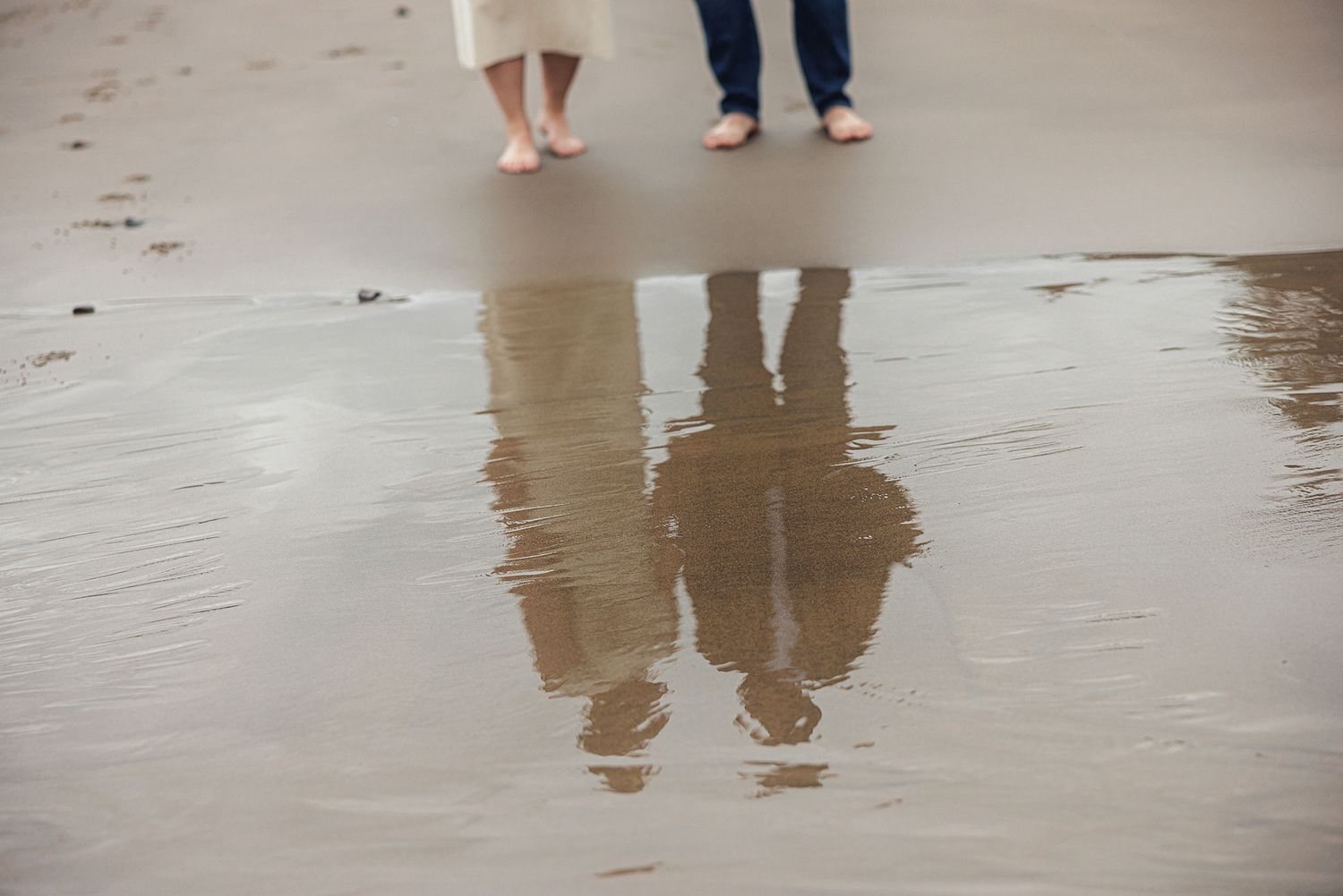 Two barefoot people standing on wet sand at the beach, with their reflections visible in a shallow pool of water.