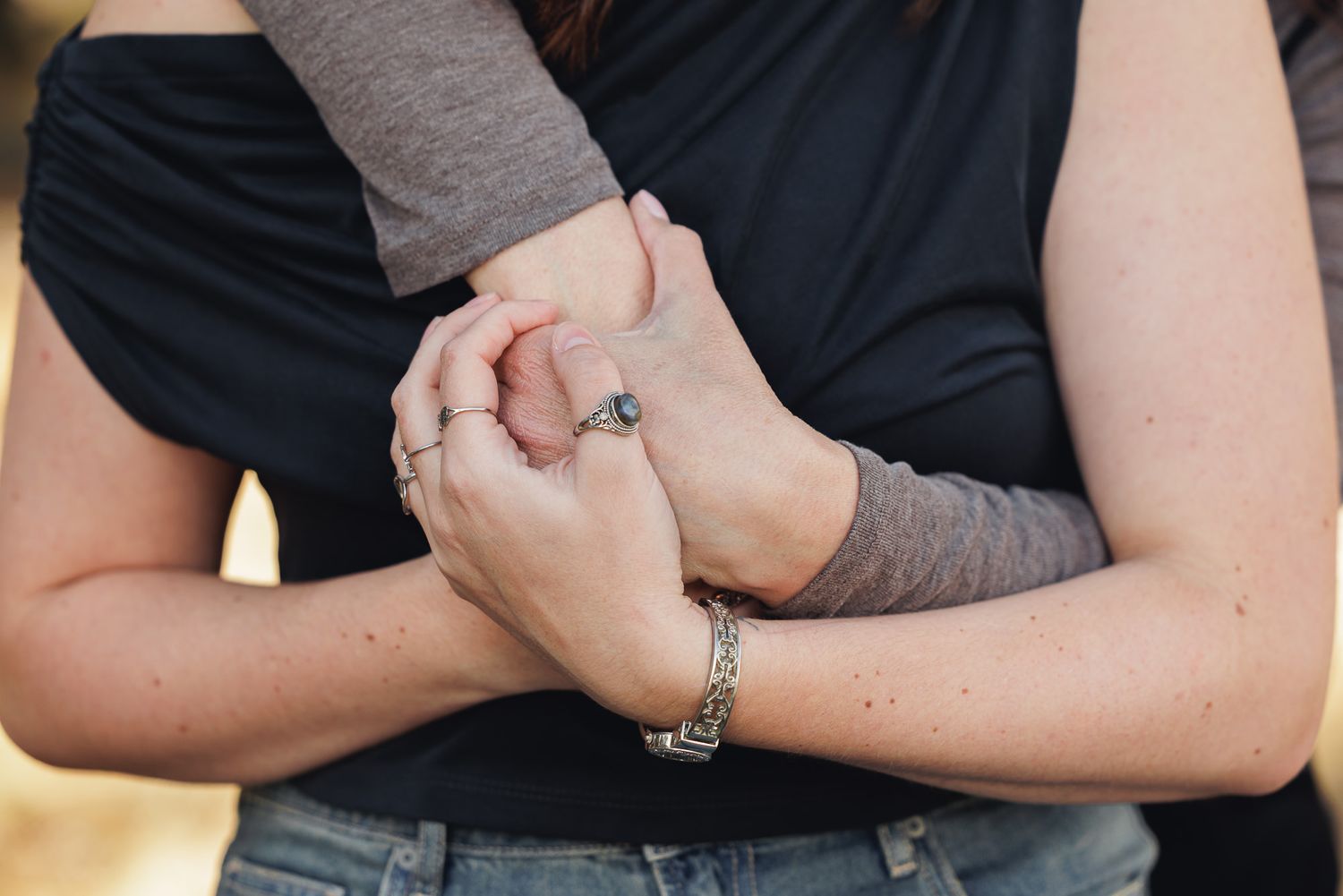 Two women holding hands affectionately, wearing bracelets and rings, in a close embrace outdoors.