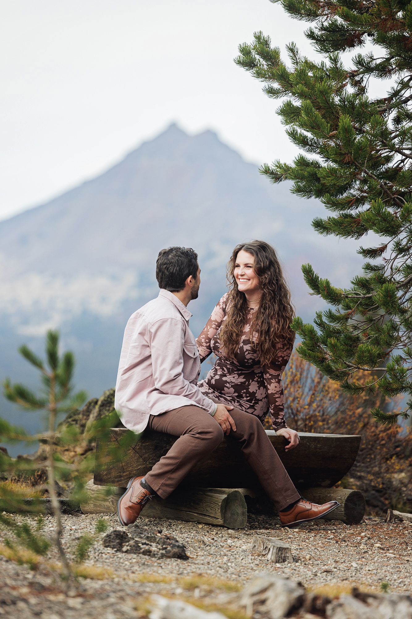 A couple sits together on a rocky mountain ledge with evergreen trees and mountain peaks in the background.