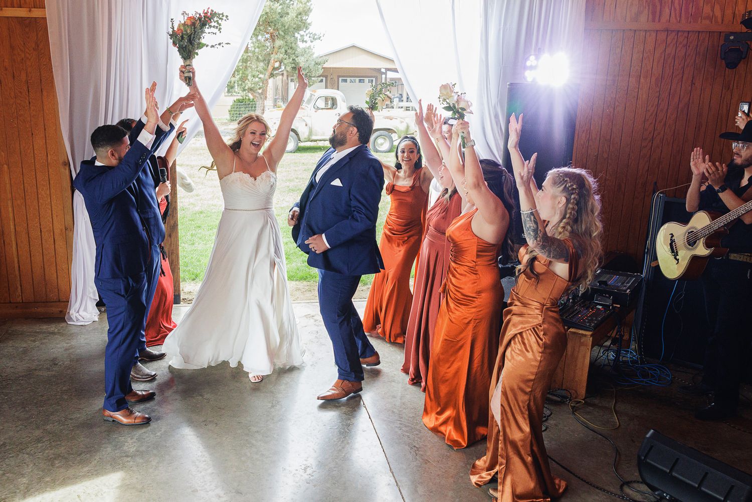 Bride and groom celebrate grand entrance at rustic wedding reception with bridesmaids in orange dresses and live band.