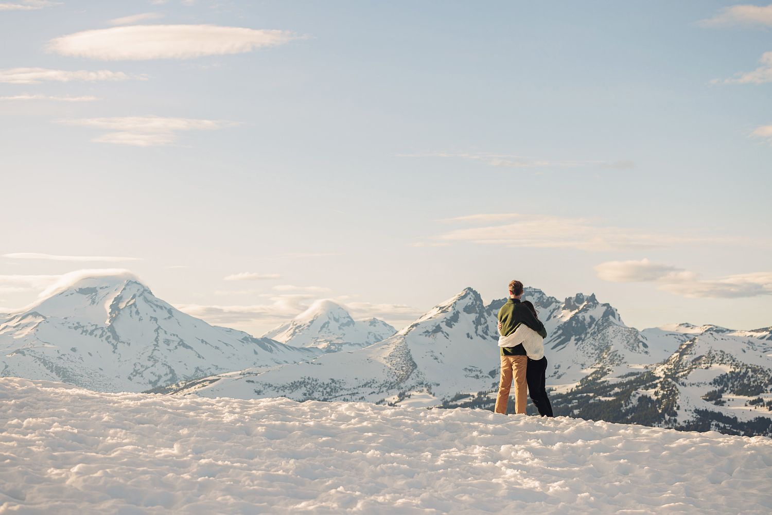 Couple embracing on a snowy mountain summit with breathtaking snow-capped peaks and soft winter sky backdrop.