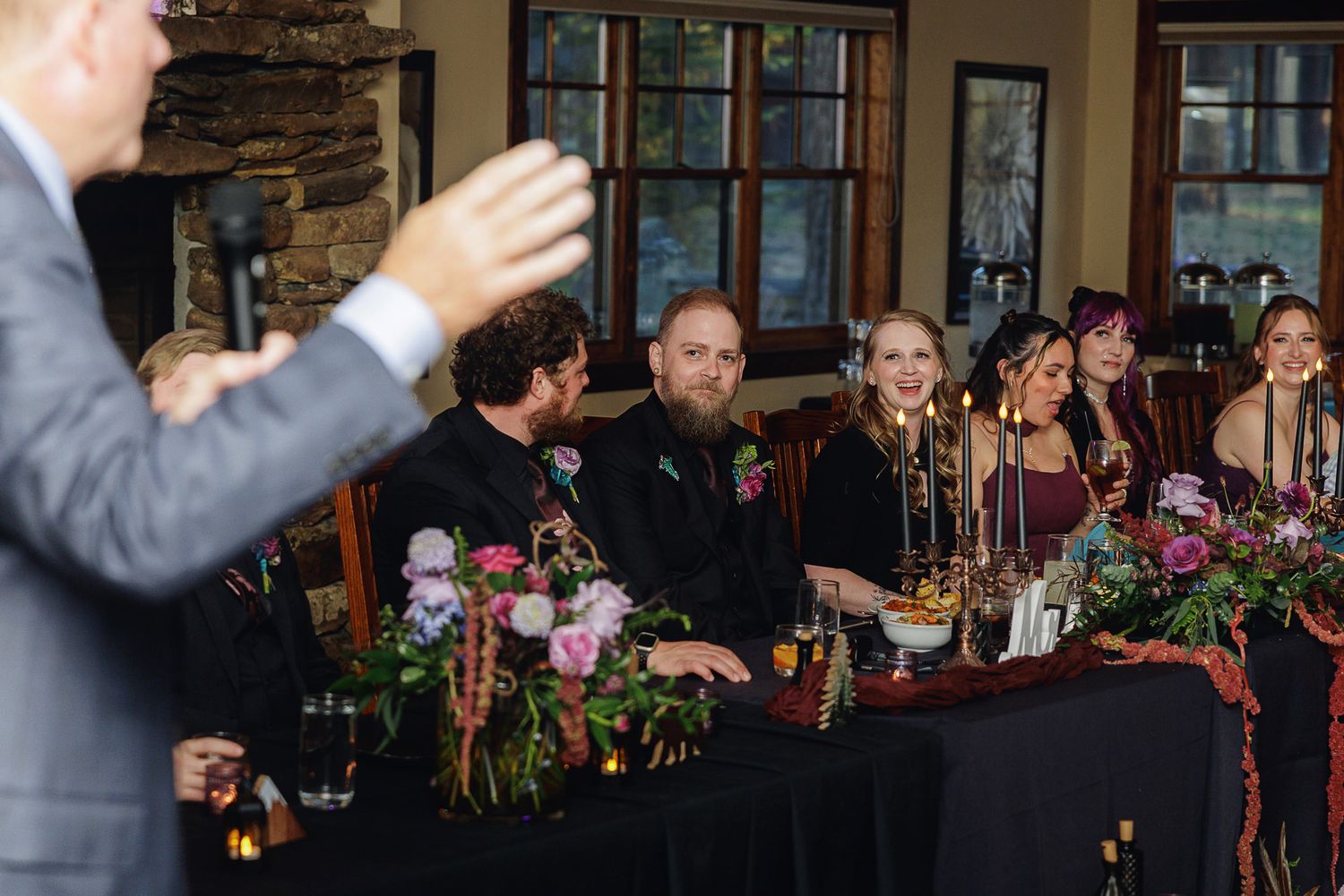 Wedding guests laughing and enjoying speeches at a rustic reception venue with floral centerpieces.