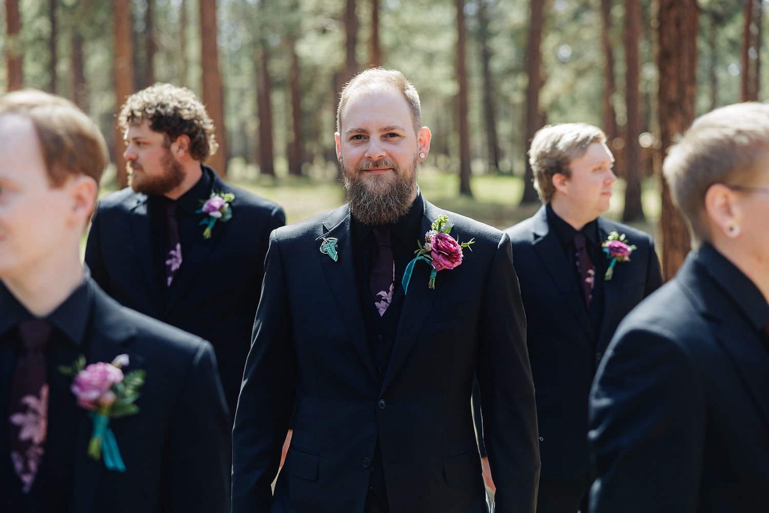 Groom with beard in black suit and floral boutonniere stands with groomsmen in matching suits among pine trees.