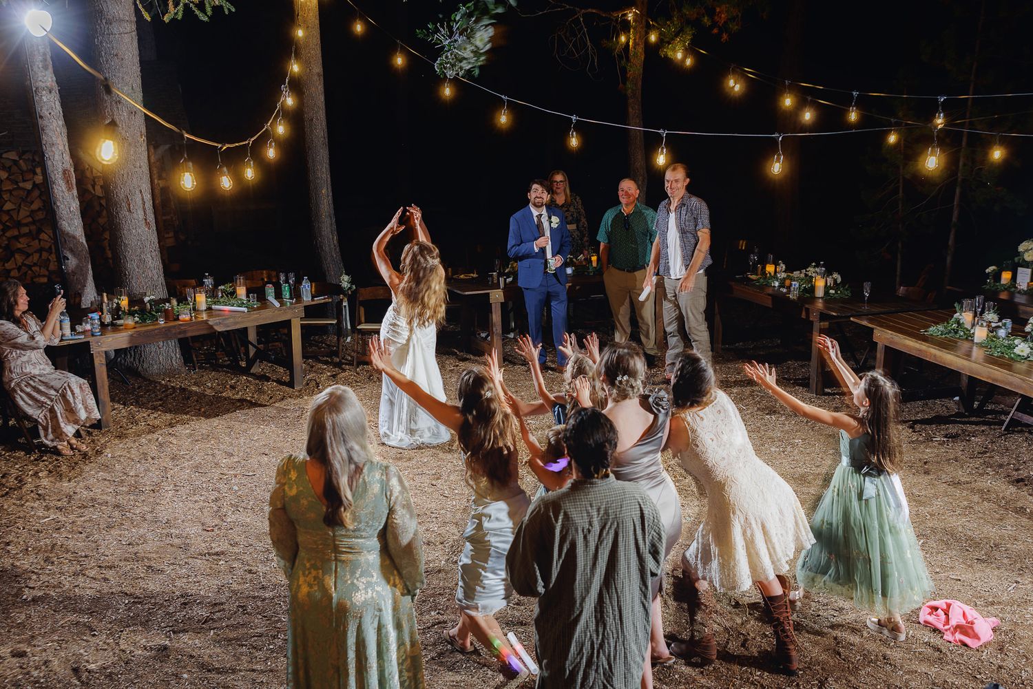 Wedding guests dance under romantic string lights at an outdoor evening reception in a wooded setting.