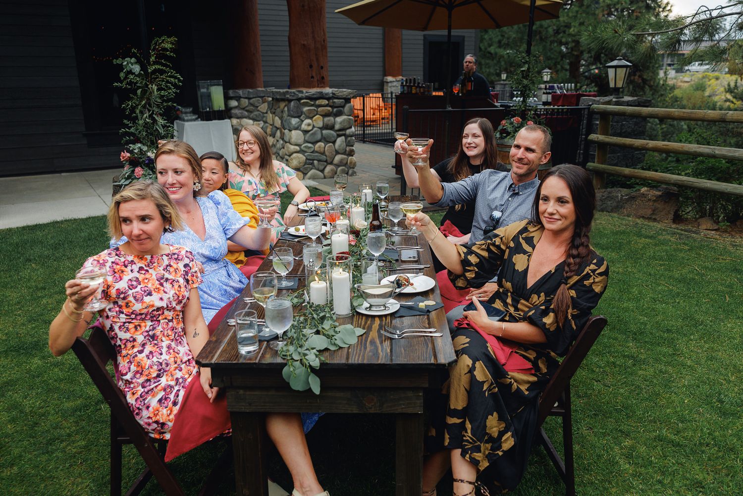 A group enjoys an outdoor dinner party at a long wooden table set up in a backyard garden setting.