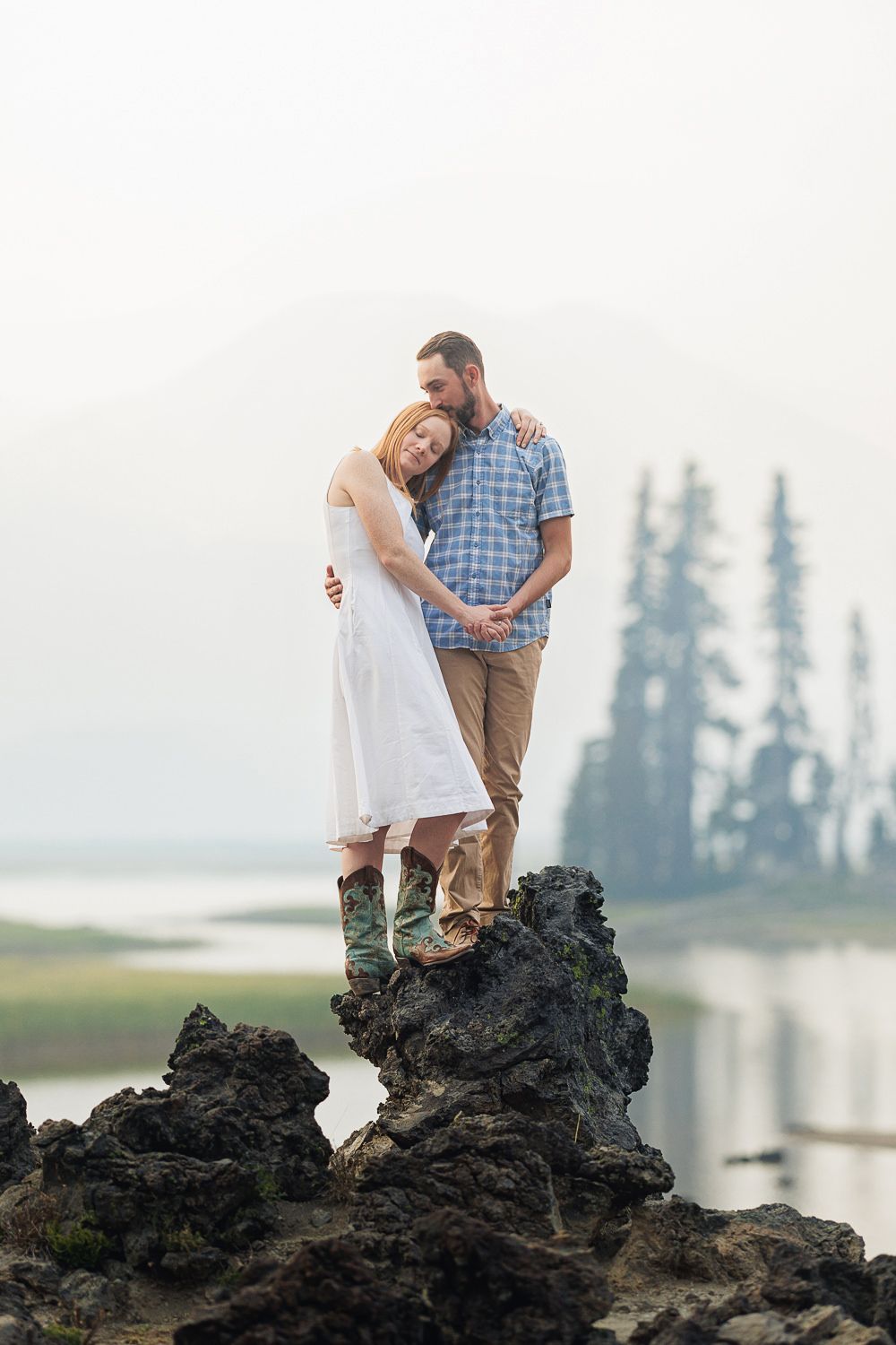 A couple stands together on a rocky outcrop overlooking a misty lake with evergreen trees in the background.