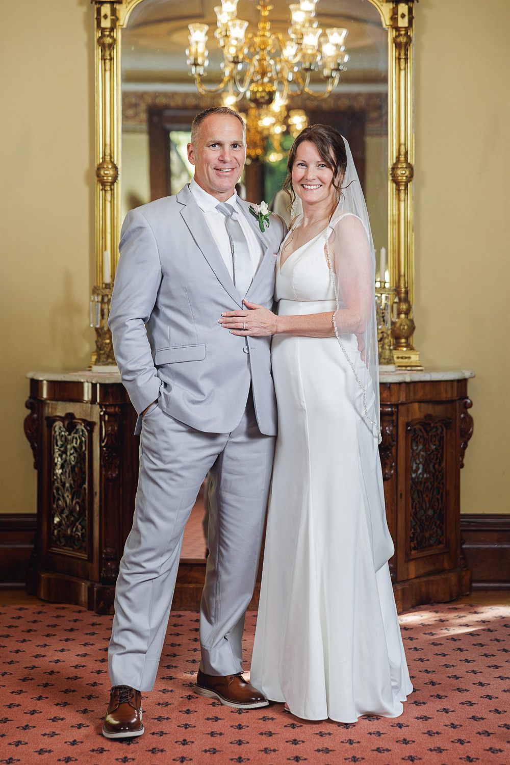 A couple in wedding attire poses together in an elegant room with chandeliers and antique furniture in multiple angles.
