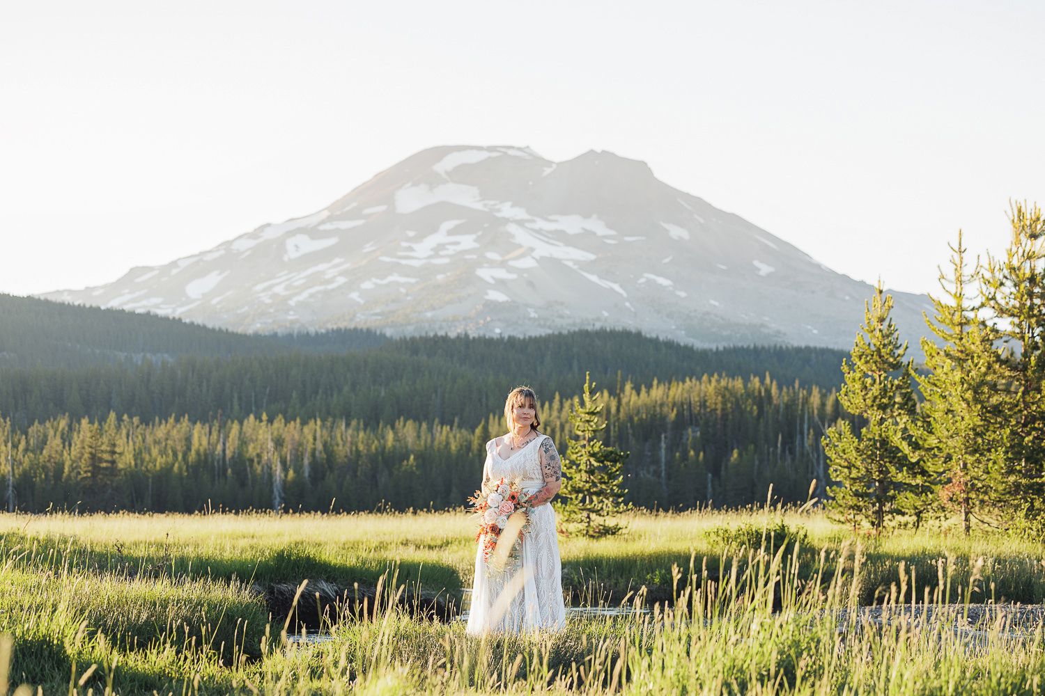 A figure in a white dress stands in a meadow with Mount Bachelor and evergreen forest in the background at sunset.