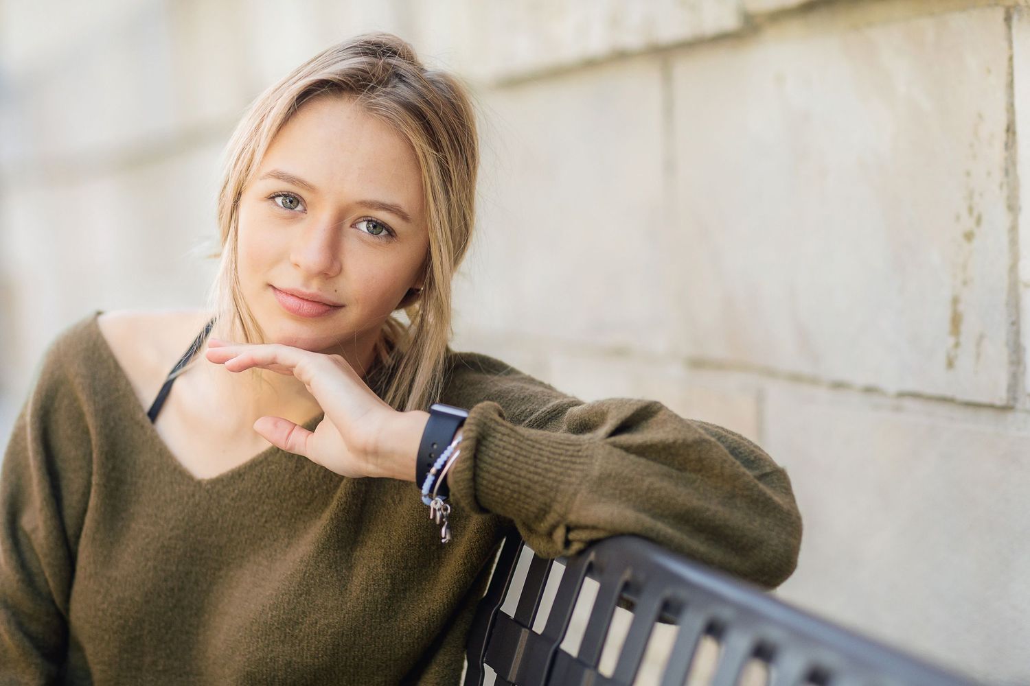 Person in olive sweater rests chin on hand while sitting against white wall in casual portrait.
