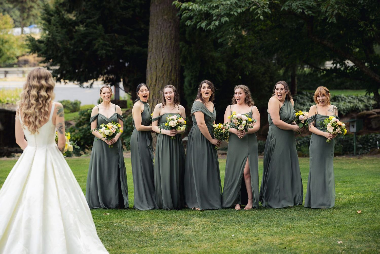 A wedding party stands on a lawn with bridesmaids in sage green dresses holding sunflower bouquets beside a bride in white.