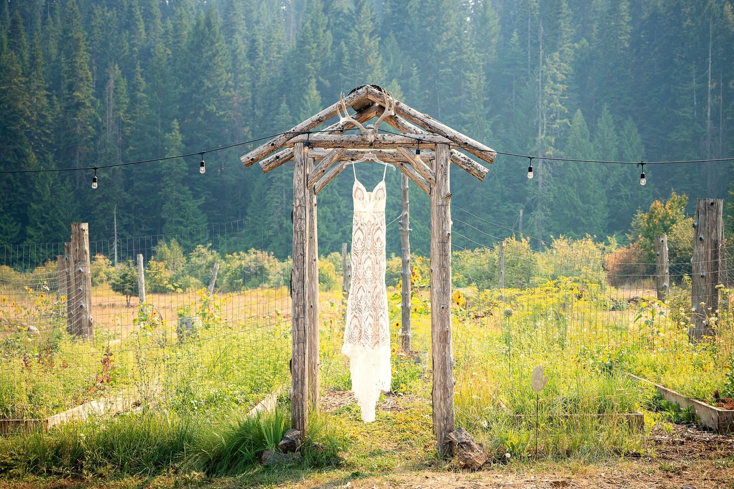 A white wedding dress hangs from a rustic wooden arch in a mountain meadow.