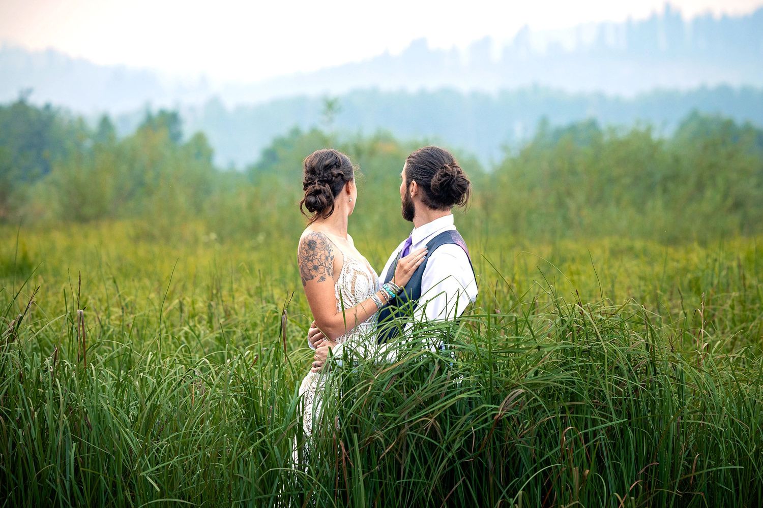 Newlyweds stand together in a field of tall grass with mountains in the background.