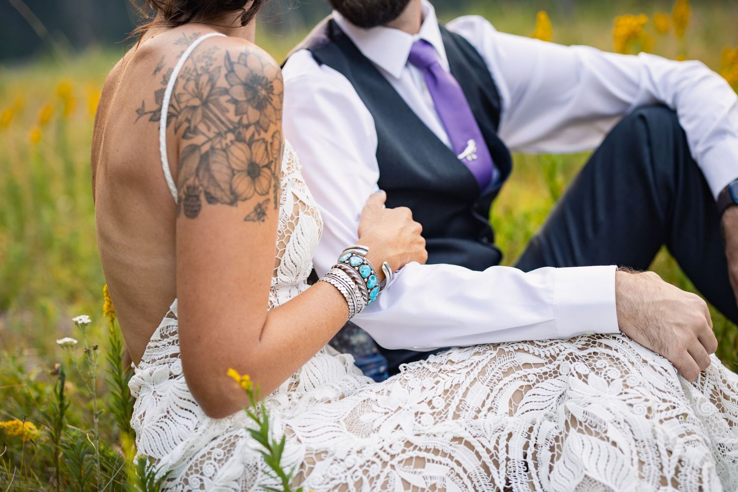 A close-up view of a white lace wedding dress and suit, with a visible tattoo on one arm sitting in a wildflower field.