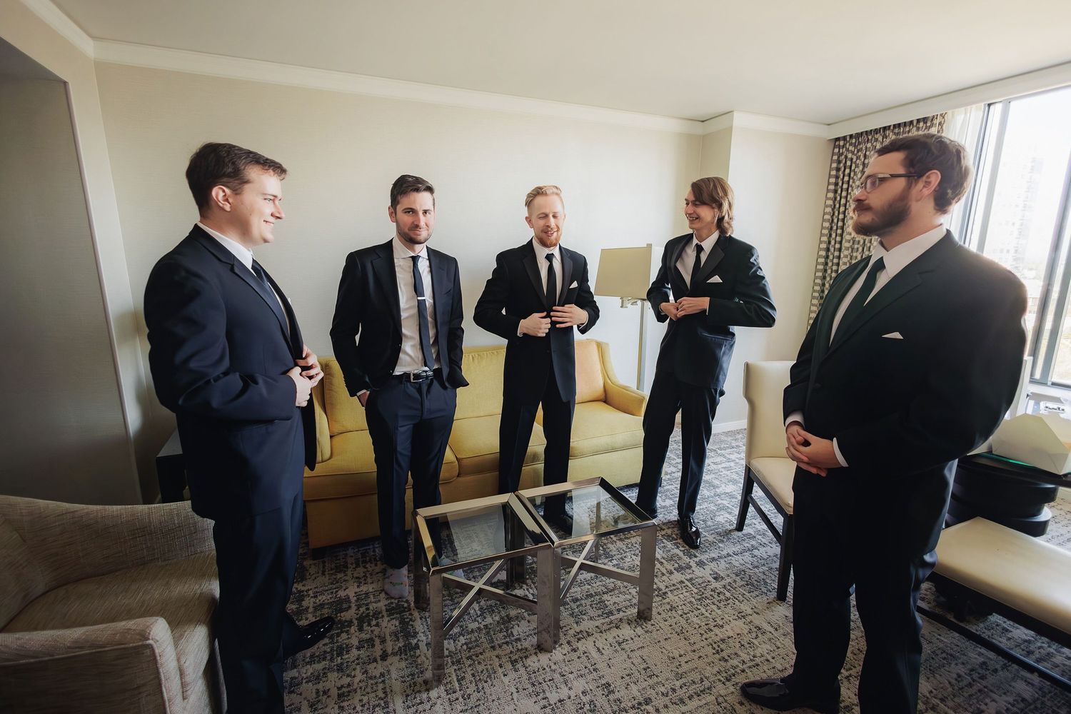 Five men in formal business suits gather for conversation in a hotel room with carpeted floor and neutral decor.