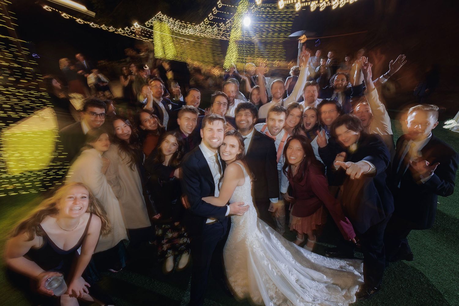 Large festive crowd celebrating at an outdoor nighttime wedding reception with string lights overhead.