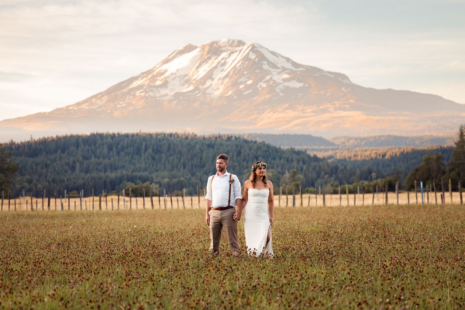 A couple stands in a golden field with Mount Adams rising majestically in the background at sunset.