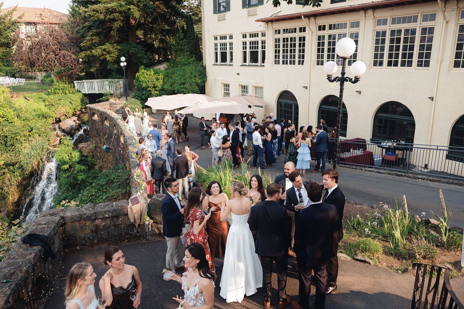 Elegant outdoor wedding reception with guests mingling near a waterfall beside a classic European building.