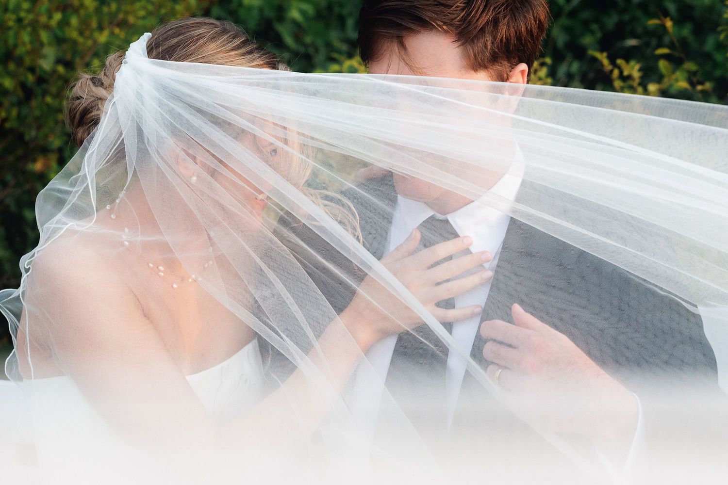 Bride and groom sharing a romantic kiss hidden beneath a flowing white wedding veil outdoors.