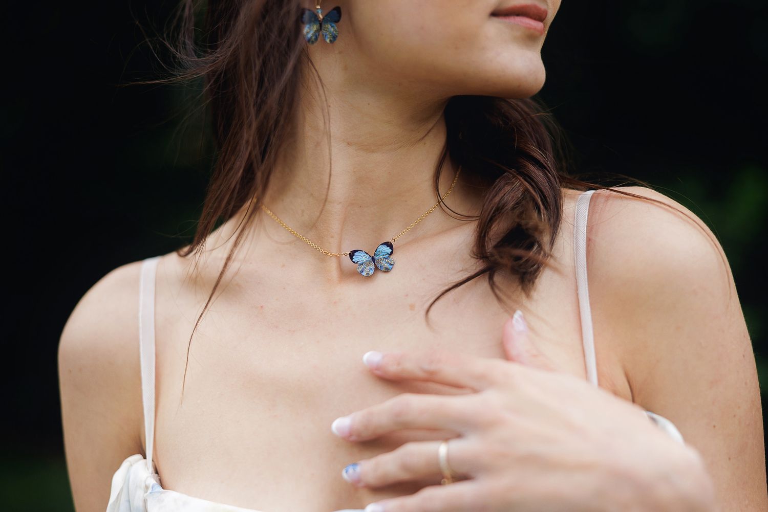 Woman wearing a blue butterfly necklace and matching earrings, with a delicate ring, in an outdoor setting.