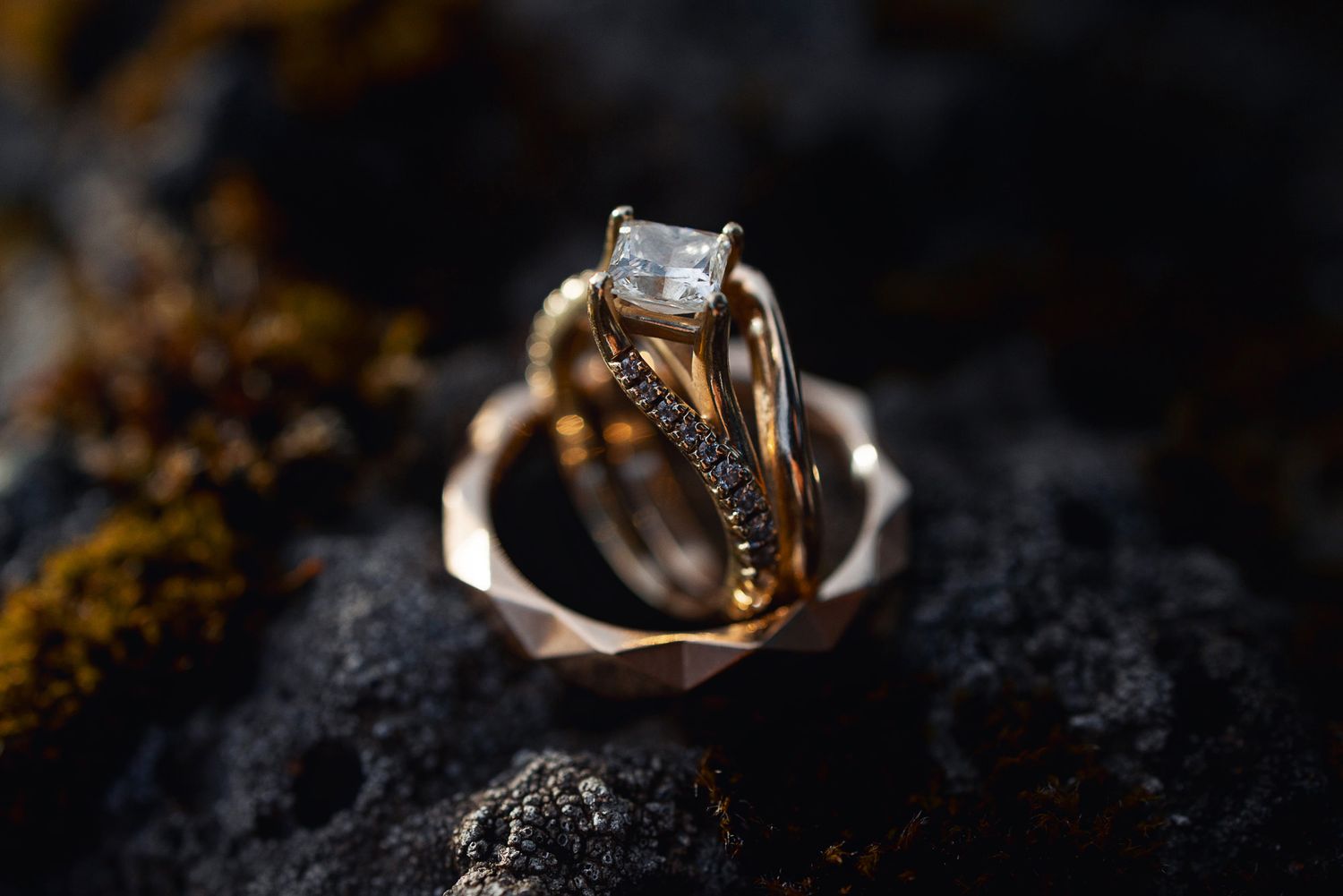 Gold engagement ring with emerald-cut diamond and matching diamond band displayed on dark rocky surface.