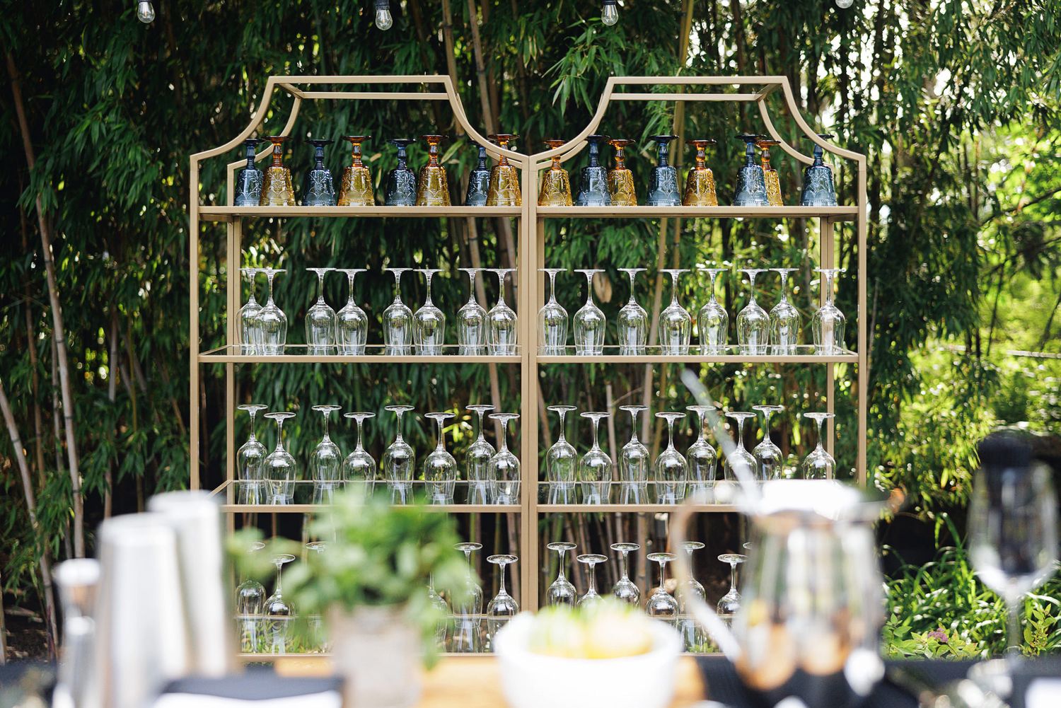 Gold bar shelf display with elegant glassware outdoors, surrounded by lush green bamboo trees at a garden event.