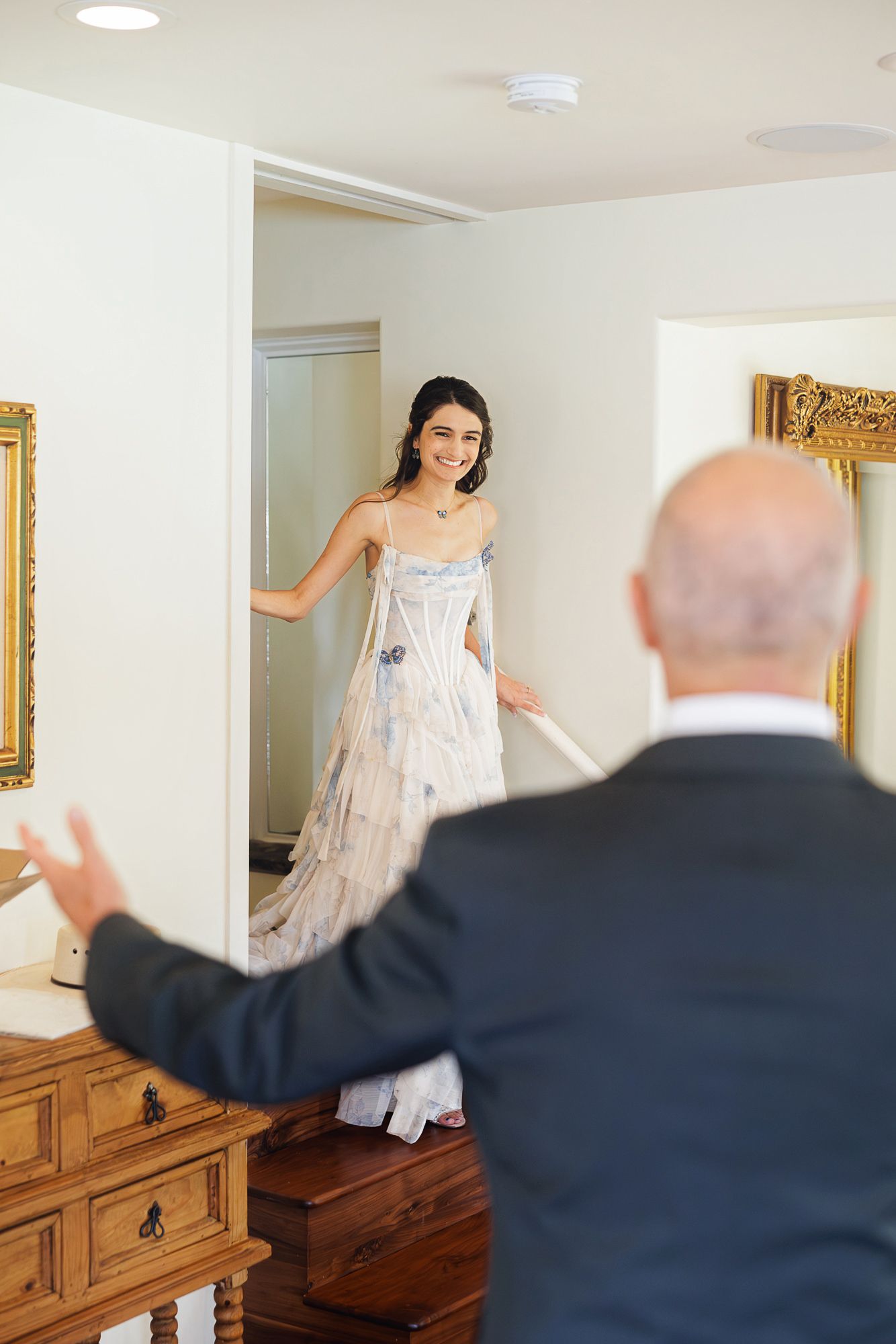 A smiling bride in a floral gown stands in a doorway as a bald man in a suit gestures toward her in a warmly decorated room.