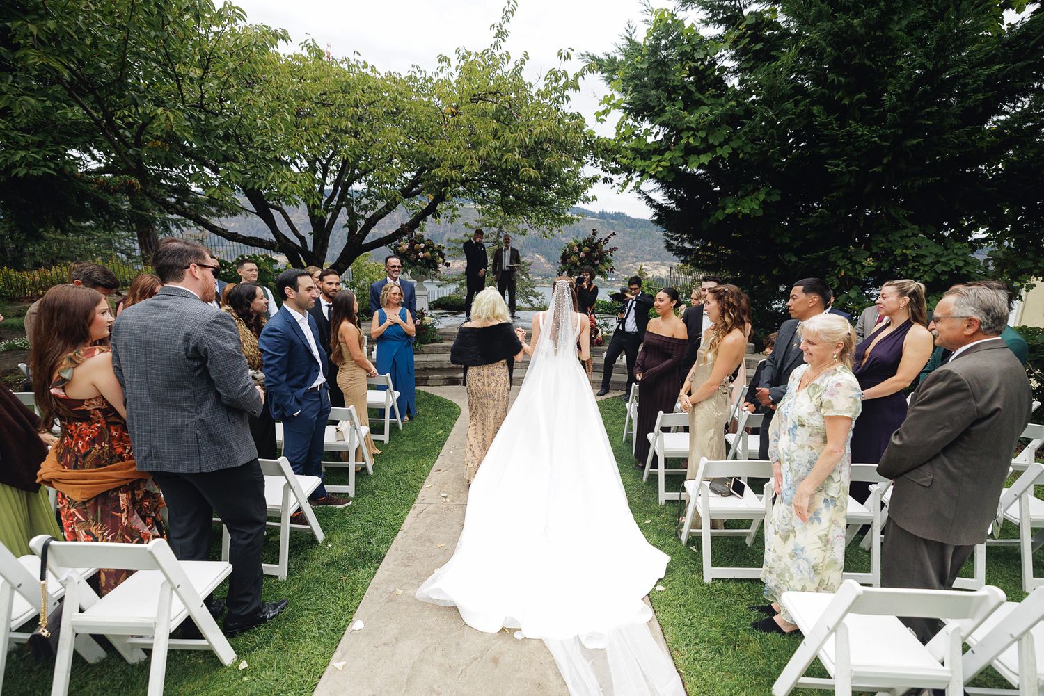 Bride walks down outdoor wedding aisle in white gown, guests seated in white chairs on green lawn.