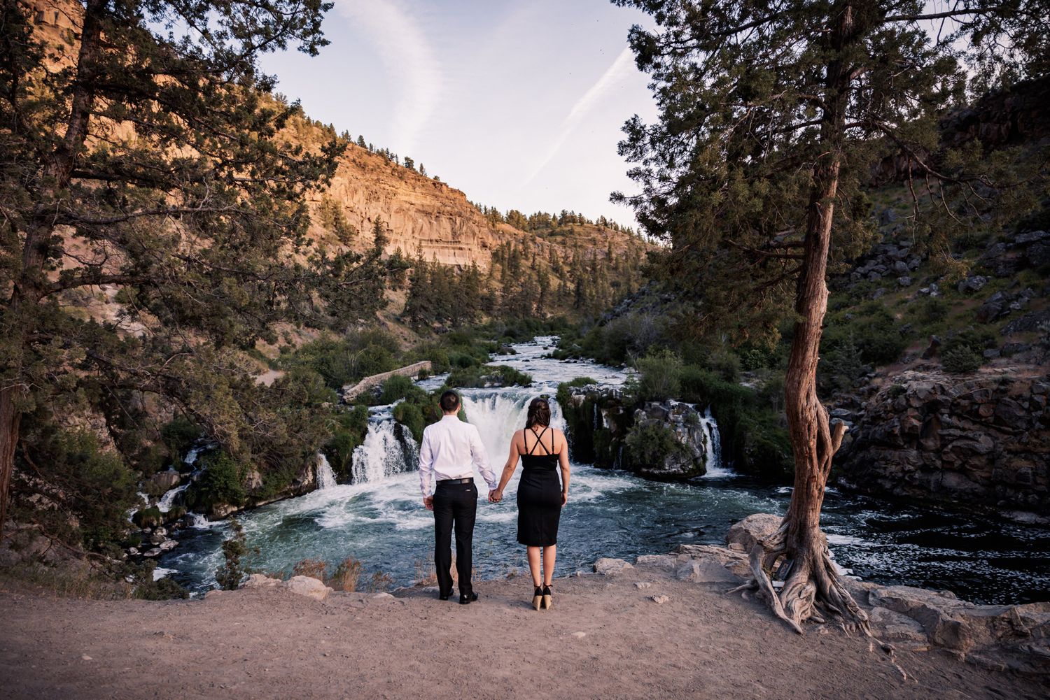 Two people stand at viewpoint overlooking Steelhead Falls waterfall in Oregon during golden hour sunset.