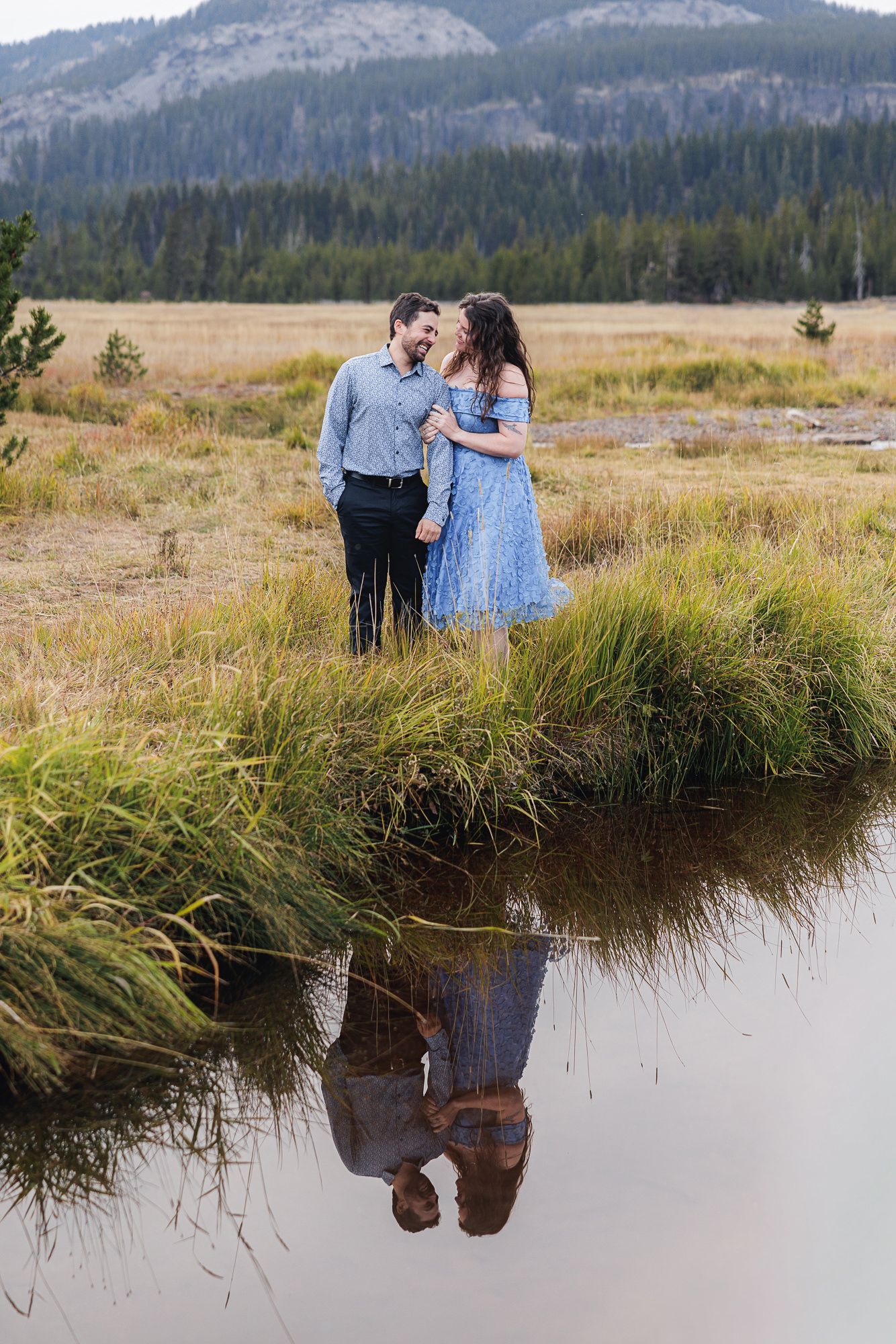 A couple shares a romantic moment by a reflective mountain stream in a scenic meadow with mountains in the background.