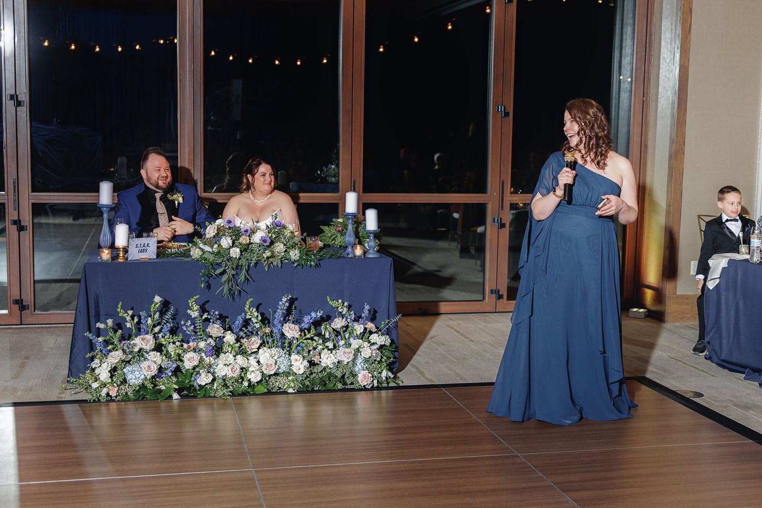Wedding reception guests in navy blue attire seated at tables with white floral centerpieces in an elegant venue.