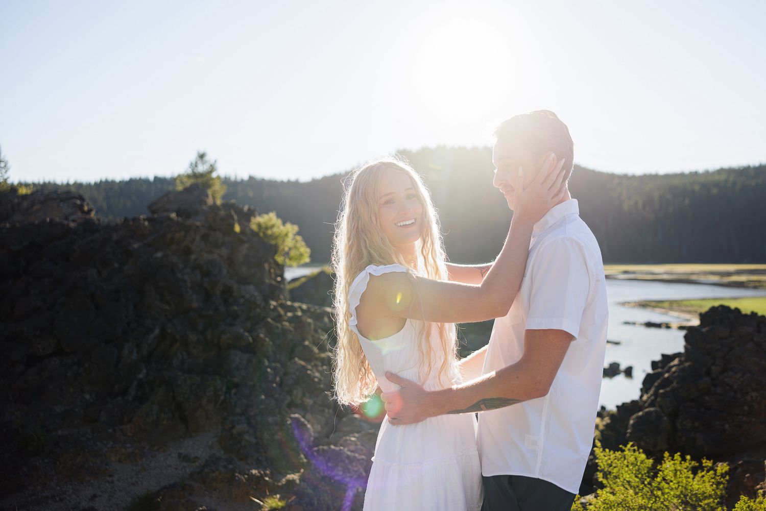 Couple embracing outdoors in bright sunlight near a lake, surrounded by lush greenery and rocky landscape.