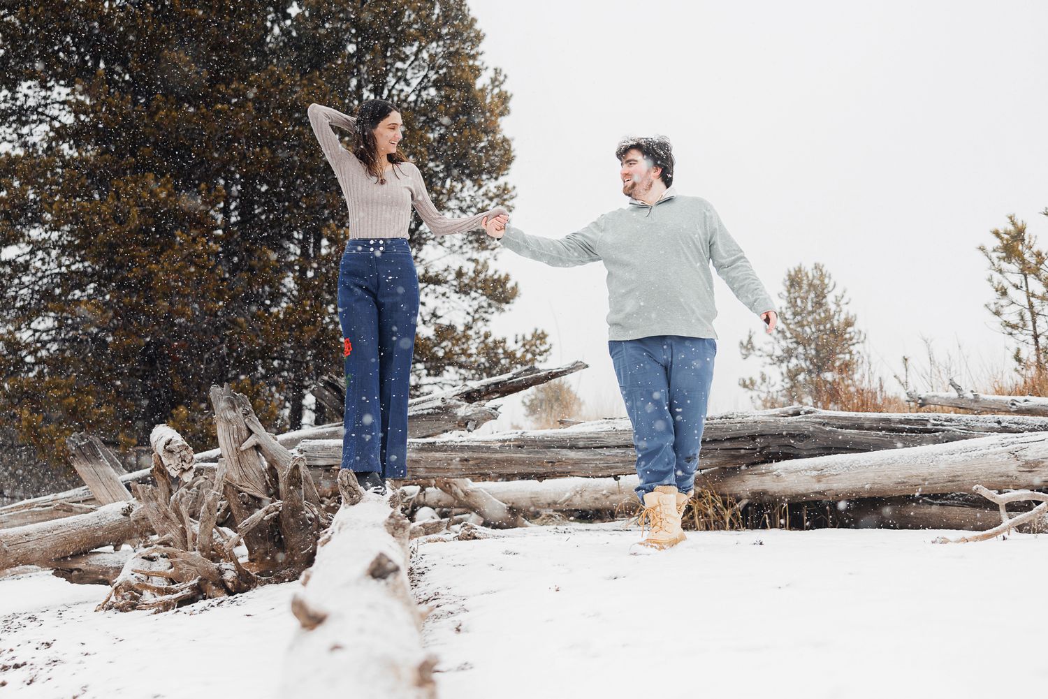 A couple playfully runs through a snowy winter landscape with pine trees in the background.