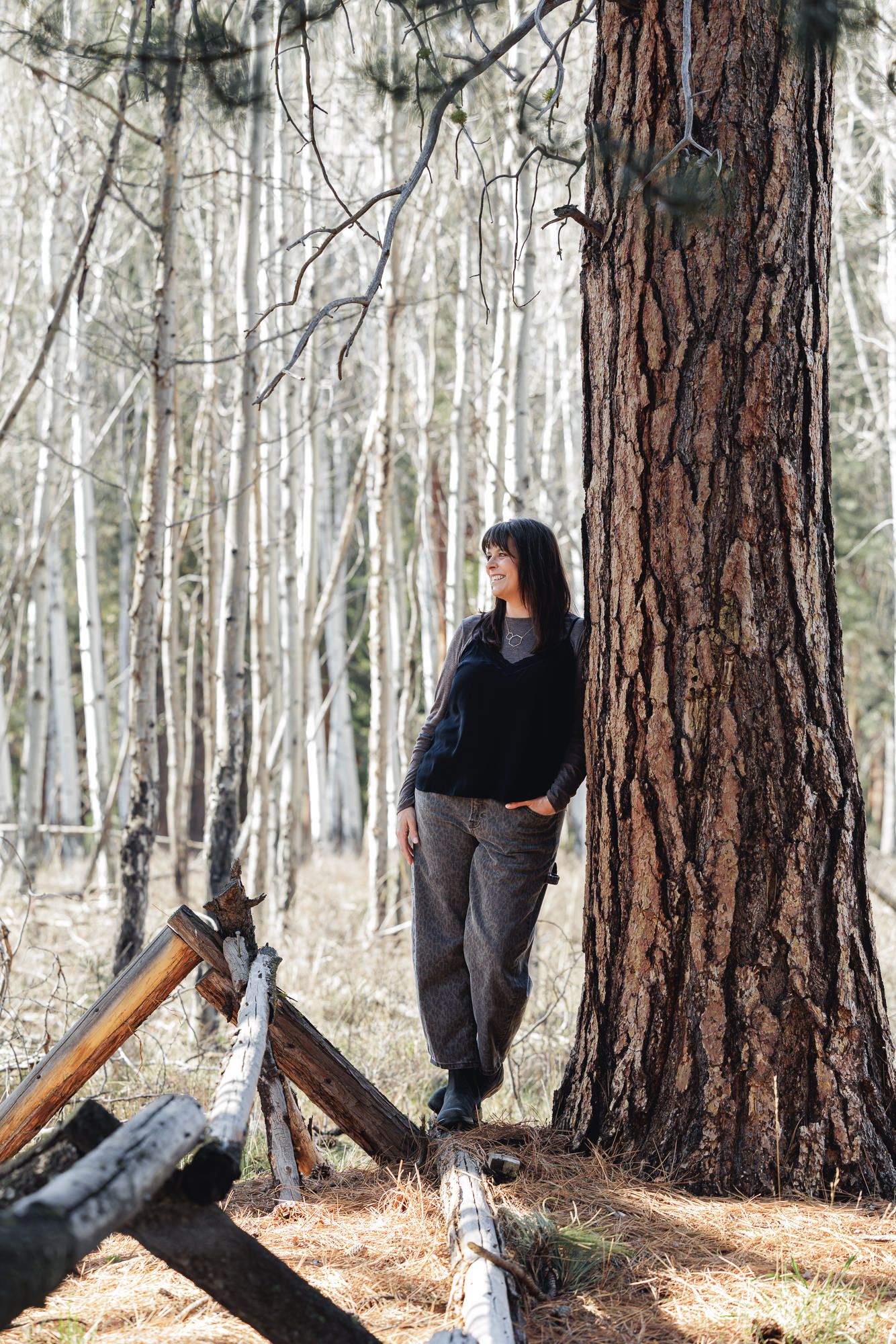 Woman in black top and gray pants leaning against a large pine tree in a sunlit forest with fallen logs.