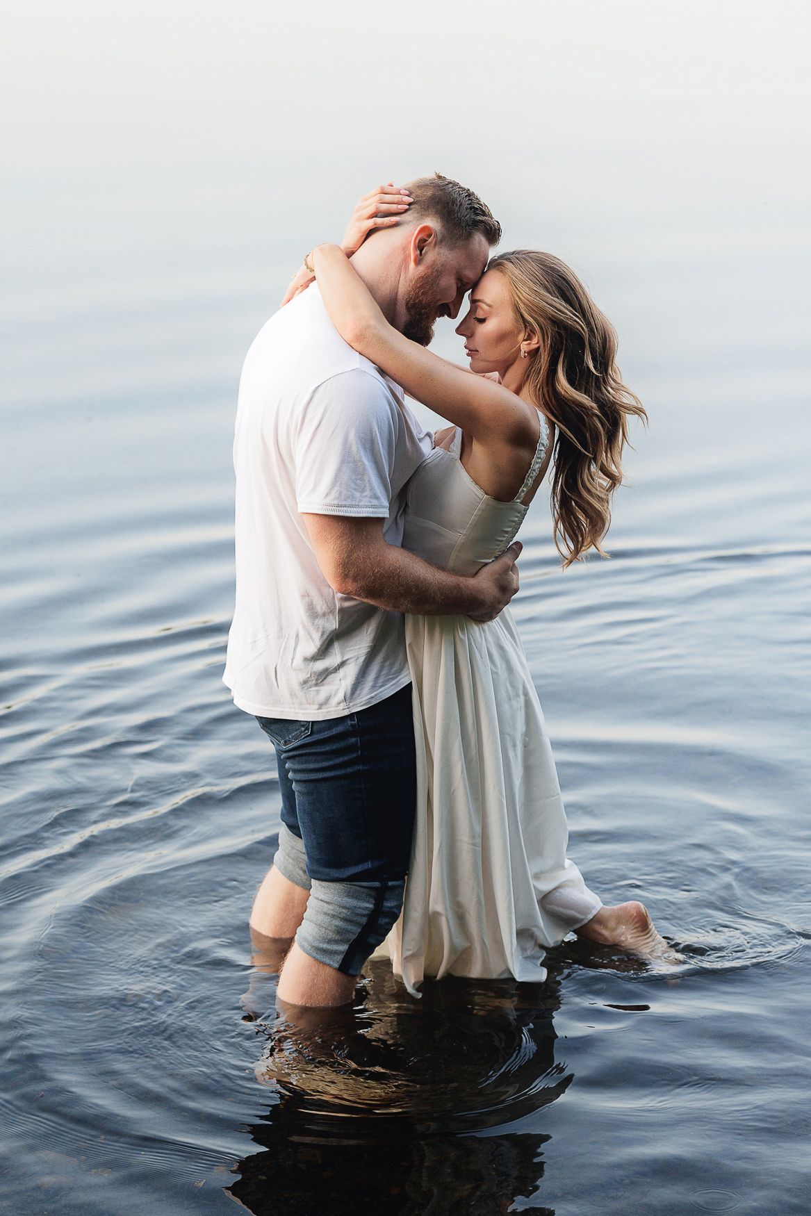 A romantic couple embraces while standing in calm lake water at sunset wearing white and denim.