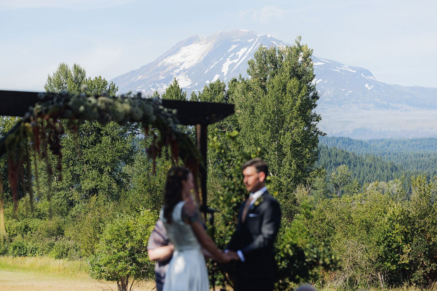 A couple stands beneath a wooden pergola with a majestic snow-capped Mount Hood rising in the background.