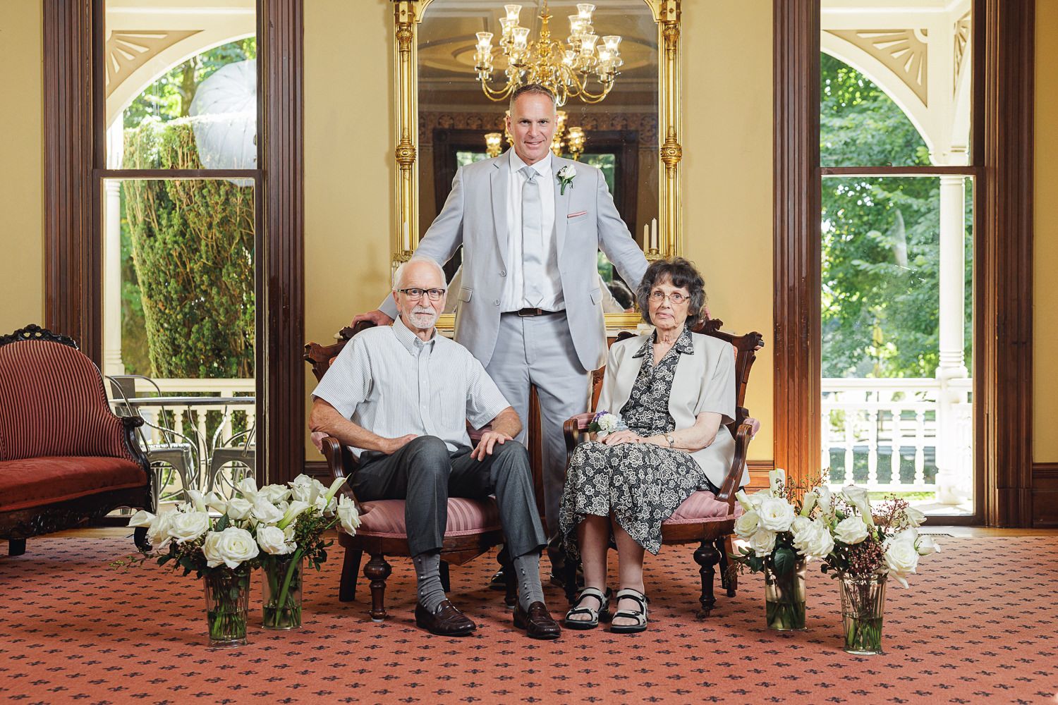 Two people in gray formal attire sit on ornate furniture in an elegant room with classical decor and floral arrangements.