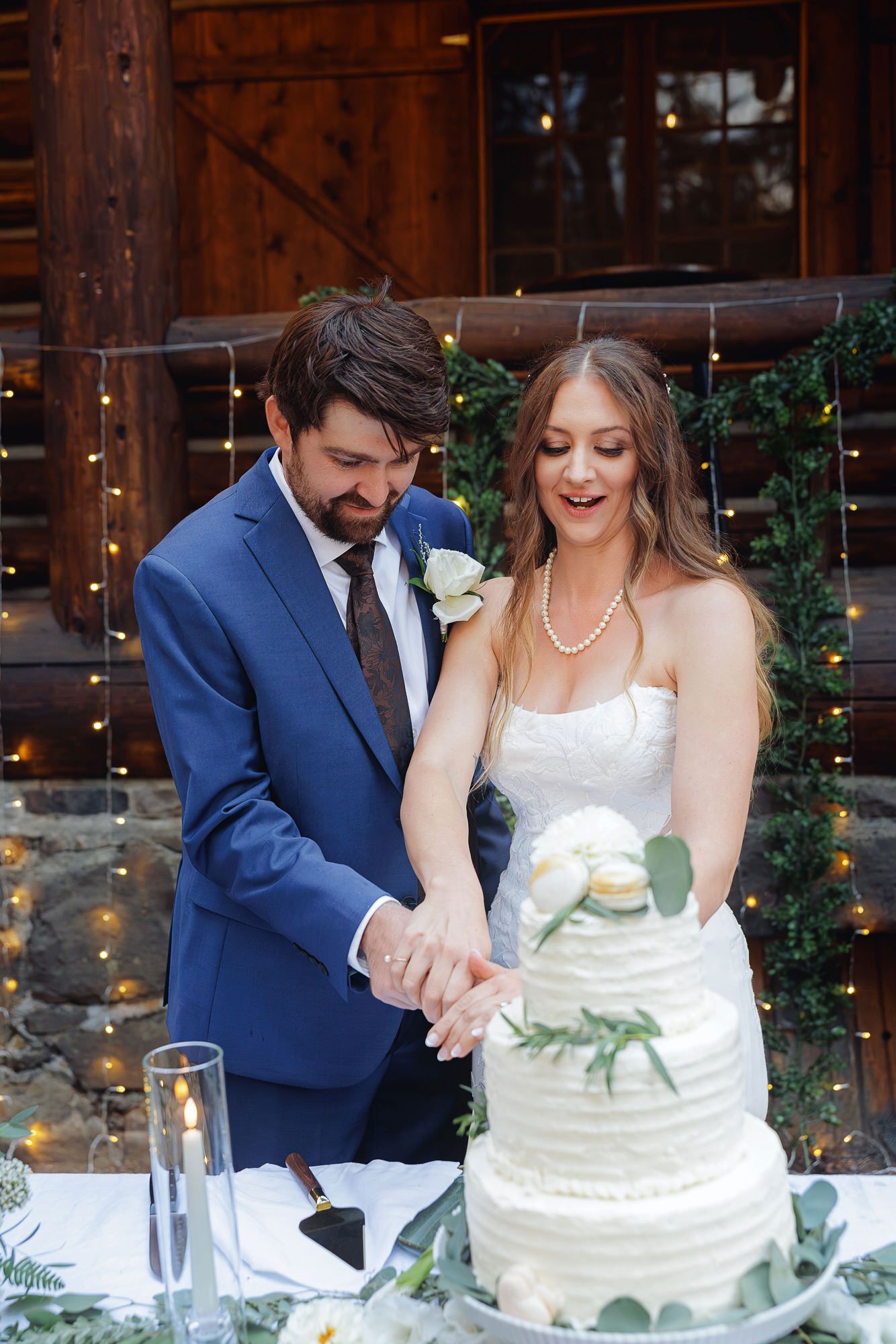 A couple in wedding attire cuts their elegant white wedding cake together at a rustic barn reception.