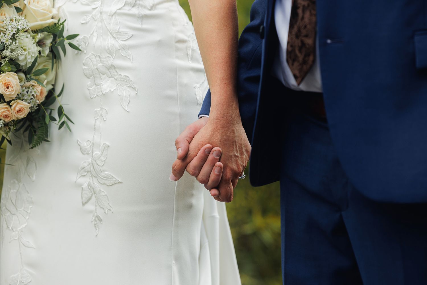 A close-up view of a couple holding hands at their wedding, with the bride wearing a white dress and holding flowers.