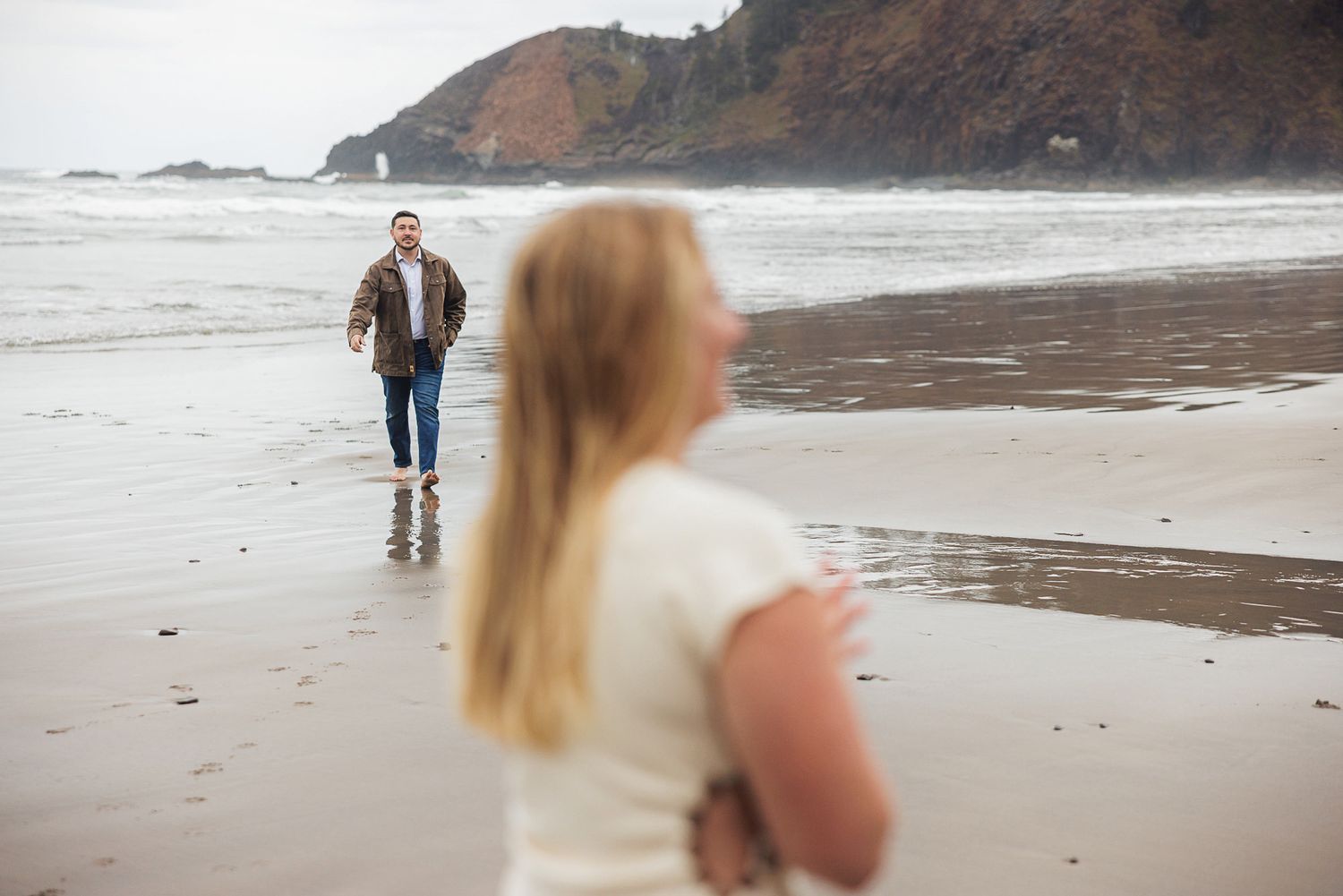 Couple on a sandy beach, woman in foreground smiling as man walks toward her with ocean waves and cliffs behind.